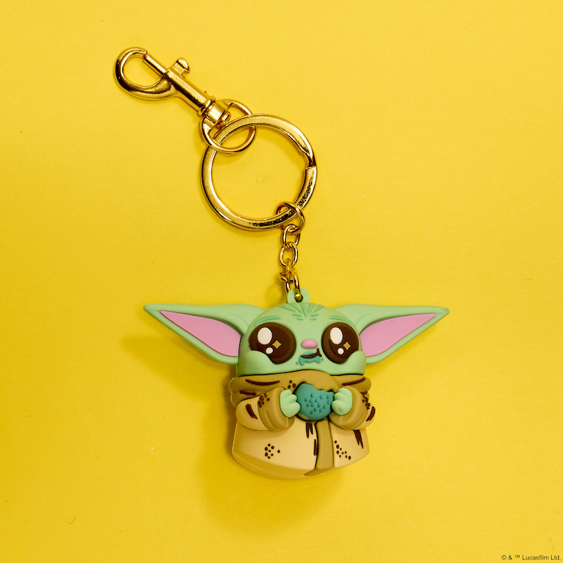3D silicone keychain in the shape of Grogu from The Mandalorian, with starry eyes and holding a half-eaten cookie. the keychain rests against a yellow background. 